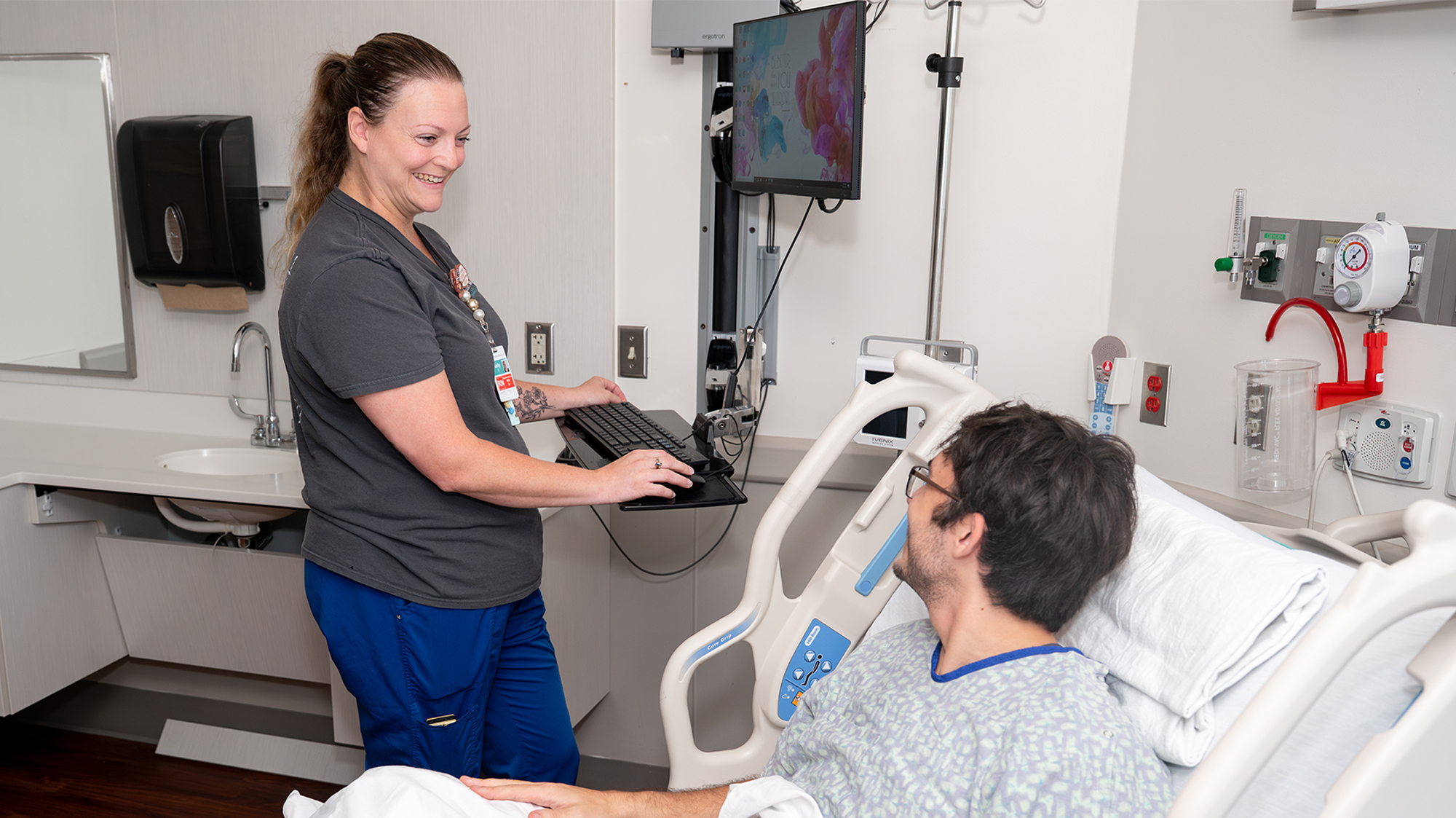 A nurse stands at a computer station in a patient room and smiles at the patient laying in the bed next to her