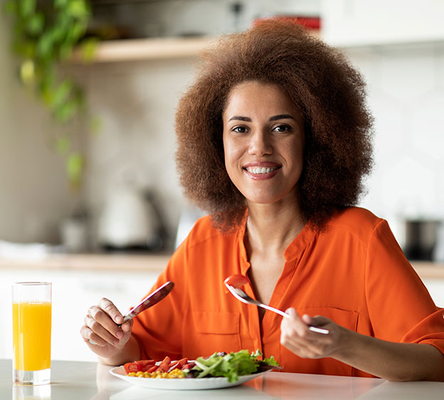 Smiling Black woman wearing an orange blouse and eating breakfast or lunch at table in kitchen