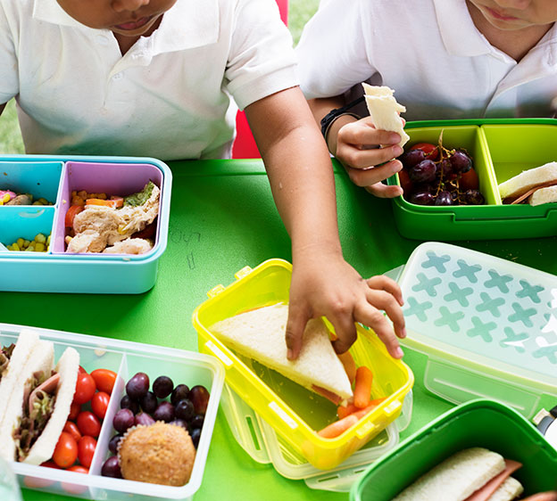 A group of two children wearing white polo shirts eating lunch at a table with lunch and bento boxes surrounding them while one of the children reaches for a half sandwich from one of the boxes