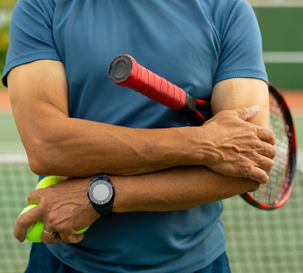 An older adult wearing blue tennis-appropriate activewear is photographed from the neck down until the upper thigh and is holding a tennis racket under their left arm