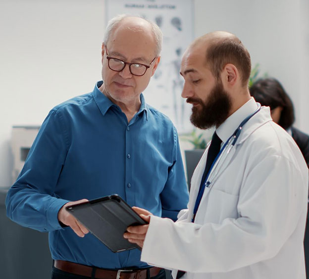 Male physician consulting with older man in hospital reception lobby looking down at a tablet together