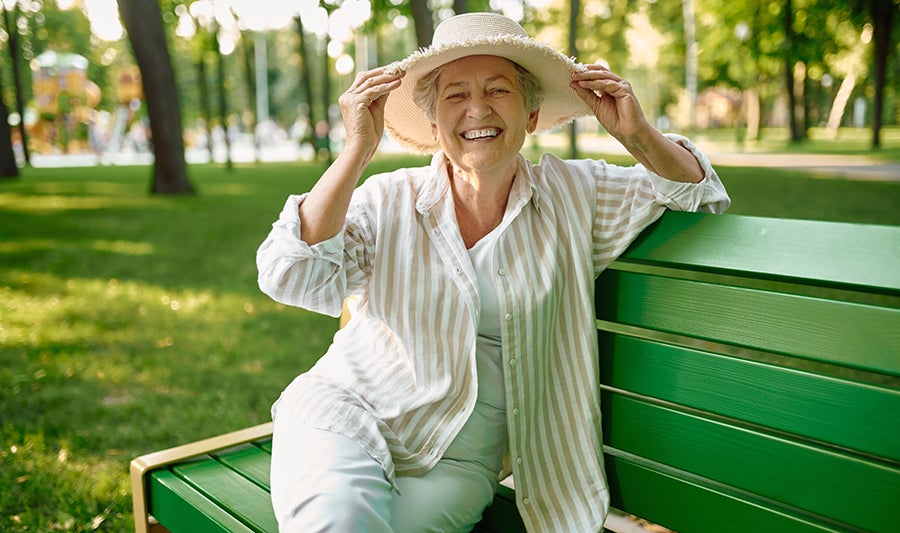 Aging woman sitting on a green bench in a park smiling at the camera while lightly grabbing her sun hat with both hands