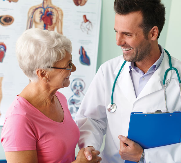 An older female patient sits on an exam table and shakes hands with a young male oncologist.