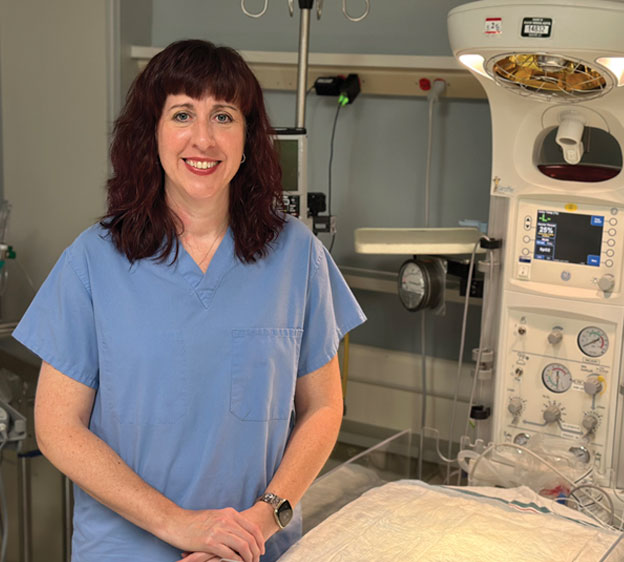 A nurse named Carrie McClure dressed in blue scrubs and standing in front of medical equipment for a photograph