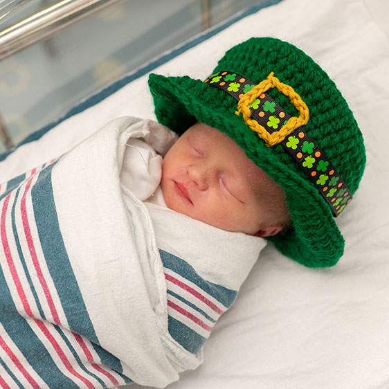 An infant in a bassinet wearing a crocheted green leprechaun hat