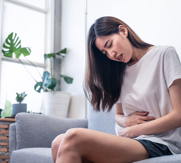 A young woman sits on a gray couch, holding her stomach in pain.