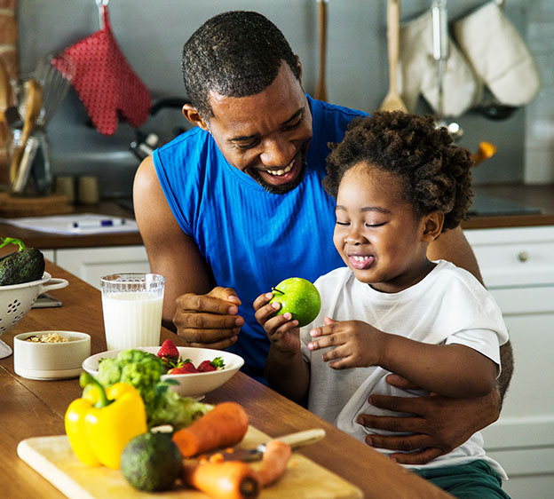 A Black man and his child sit at a kitchen counter preparing healthy foods.