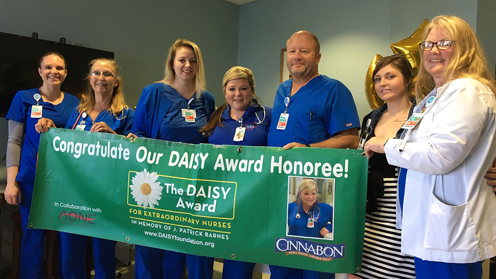 A group of nurses in blue scrubs and other medical workers photographed holding a DAISY award banner