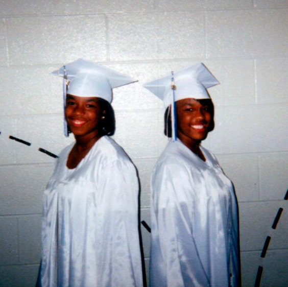 A graduation photo of the Lundy twins, who stand back to back wearing white graduation caps and gowns and smiling at the camera