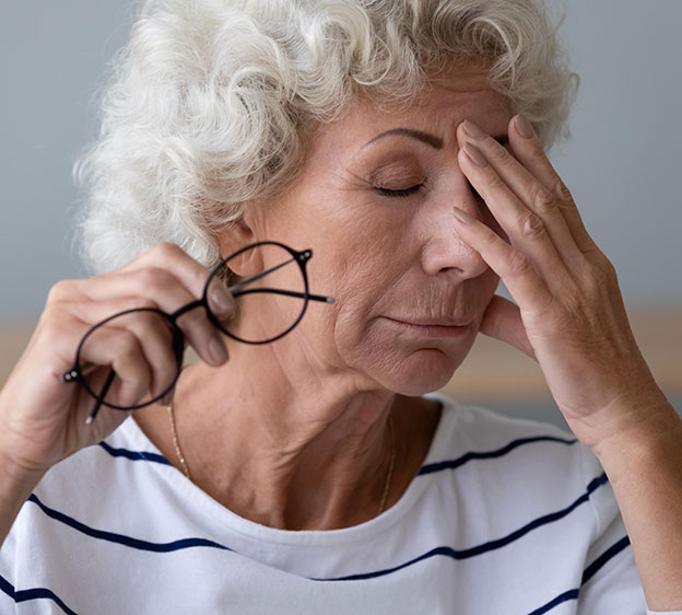 An older woman places one hand on her forehead as if in pain, while holding a pair of glasses with the other.