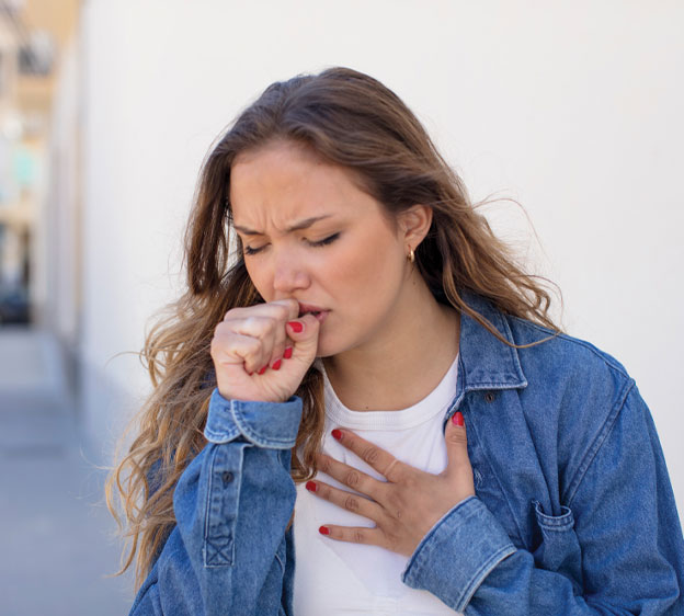 Woman in chambray shirt holding chest with one hand and holding the other hand to her mouth to stop a cough