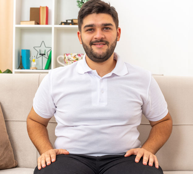 A young man wearing a white polo shirt and black pants sits on a couch and smiles at the camera.