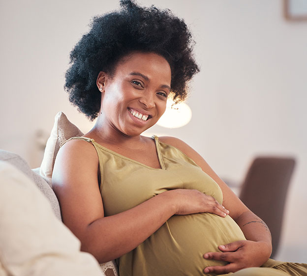 A Black pregnant woman wearing a tan sundress sits on a couch and smiles at the camera.