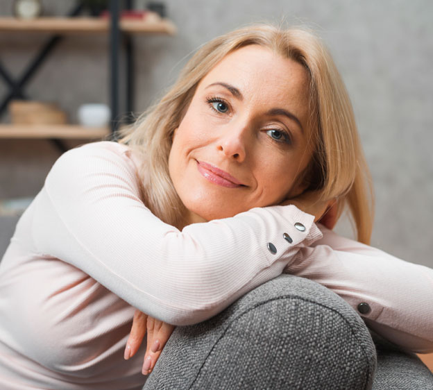 Middle-age blonde woman reclines on a gray couch and looks at the camera.