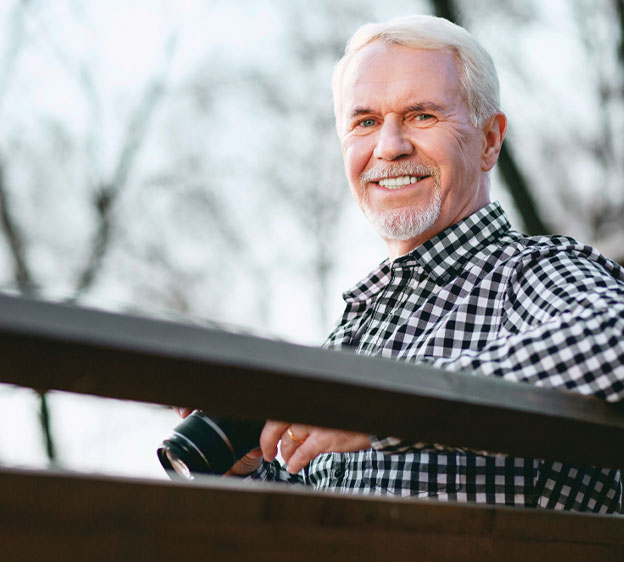 Older man standing alongside a bridge railing holding a camera and smiling.