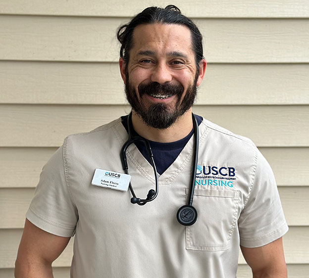 A smiling Adam Flores, a nurse in brown scrubs, poses for the camera with his name tag visible.