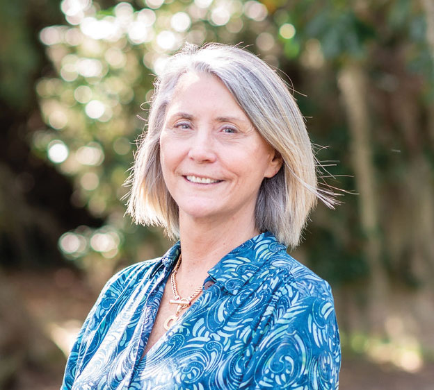 A blond woman named Roberta DeLoach in a blue dress standing and smiling toward the camera with trees in the background