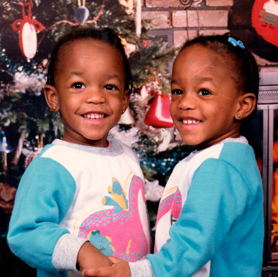 A Christmas photo of the Lundy twins as toddlers. They stand in front of a decorated tree holding hands and smiling at the camera.