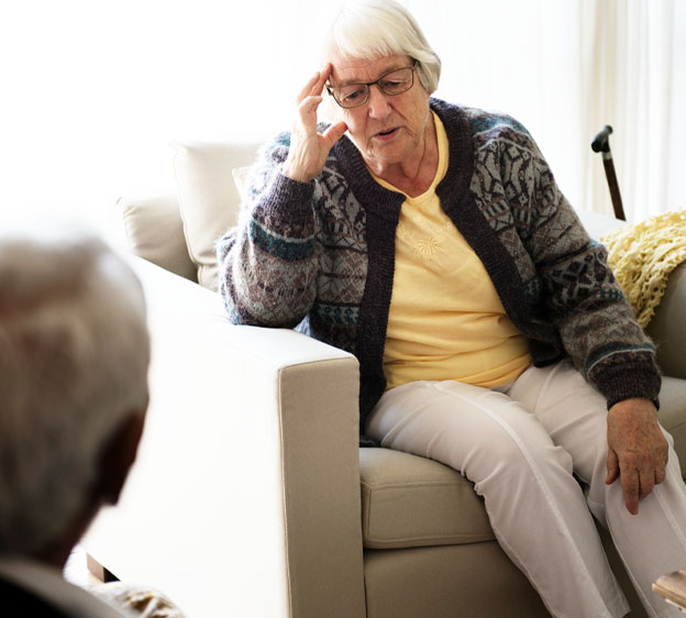 An older woman holds her head in pain as she sits on a beige armchair.