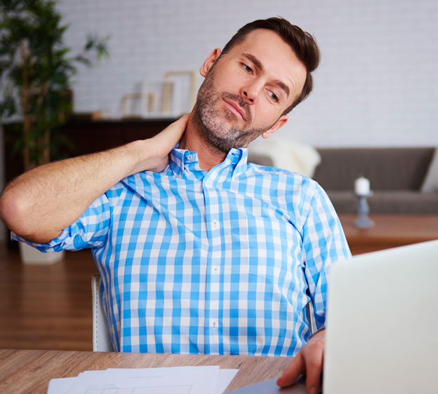 A young man sits at a desk looking down at a laptop and holding his neck in pain.