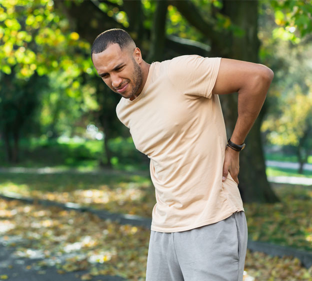 A younger man wearing athletic clothes and standing outdoors holds his back in pain.