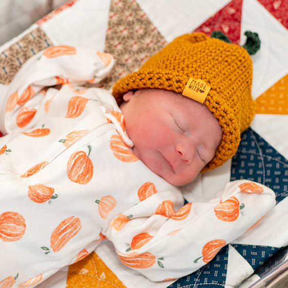 An infant in a bassinet wearing a crocheted orange Halloween hat