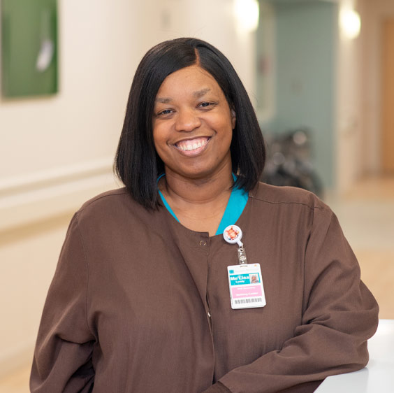 A staged photo of LeLe Lundy who smiles at the camera and wears a brown jacket over teal scrubs