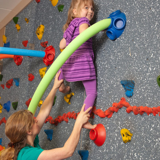 A young girl climbs up a climbing wall while a Beaufort Memorial pediatric occupational therapist supports her below