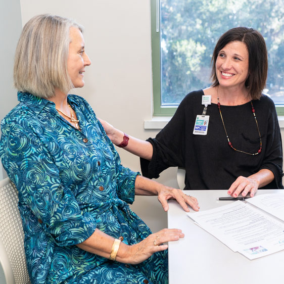 A blond woman named Roberta DeLoach in a blue dress sitting at a table and smiling toward a medical profeessional wearing a badge