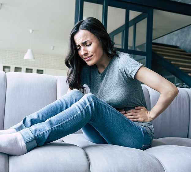 A young woman sits on a gray couch holding her abdomen in pain.