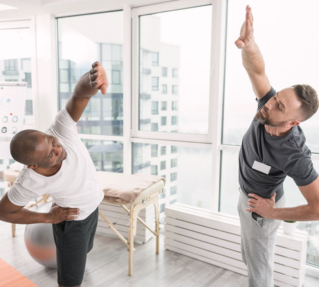 A male physical therapist works with a male patient. Both stand and stretch as they stand in a bright room with walls of windows.