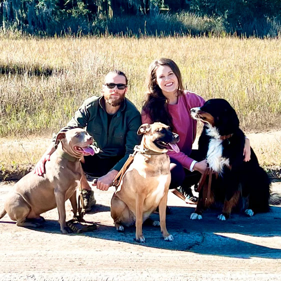 Tara Kay and her fiancé, Michael, sit on the ground outdoors with their three large dogs