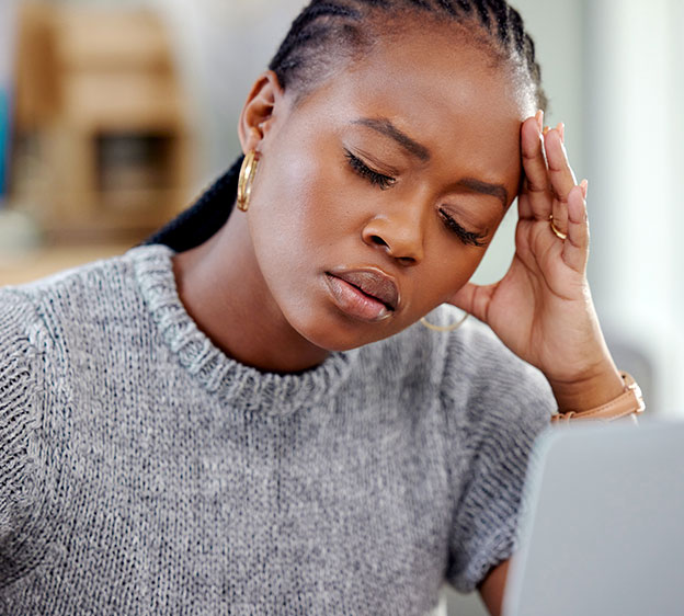 Young Black woman sitting in front of a laptop with her eyes closed, holding her head as though she has a headache