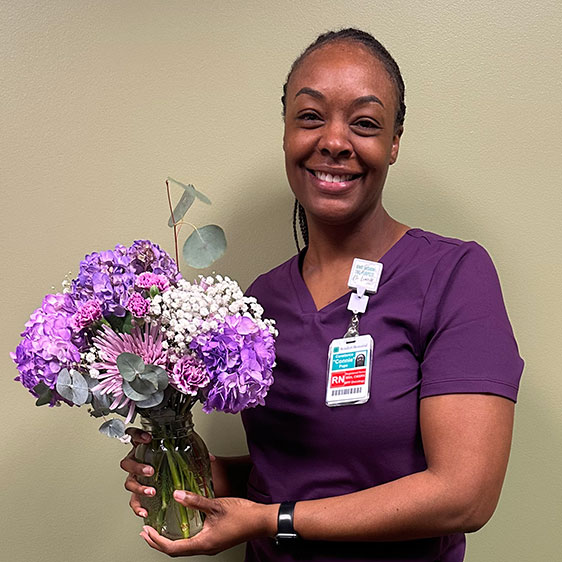 Constance (Connie) Pope wearing purple scrubs and her Beaufort Memorial badge, holding a glass vase with purple and white flowers, and smiling for a camera