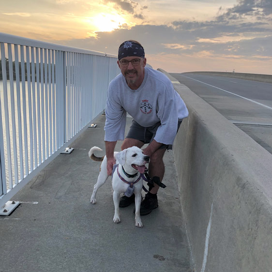 Beaufort Memorial Pharmacy Services Director Tim McCall kneels on a boardwalk with a white dog. The sun sets over the ocean in the background.