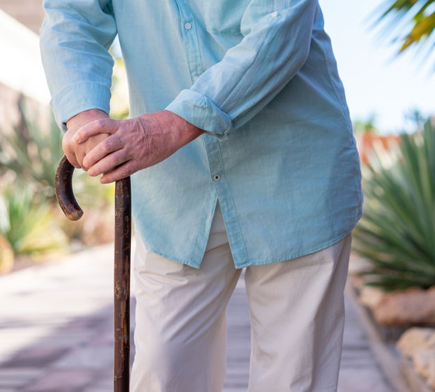 An older adult wearing a denim shirt and khakis leans on a wooden cane.