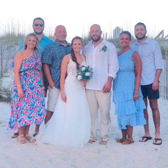 seven adults standing on the sand by a beach, two of the adults are in a wedding dress and suit