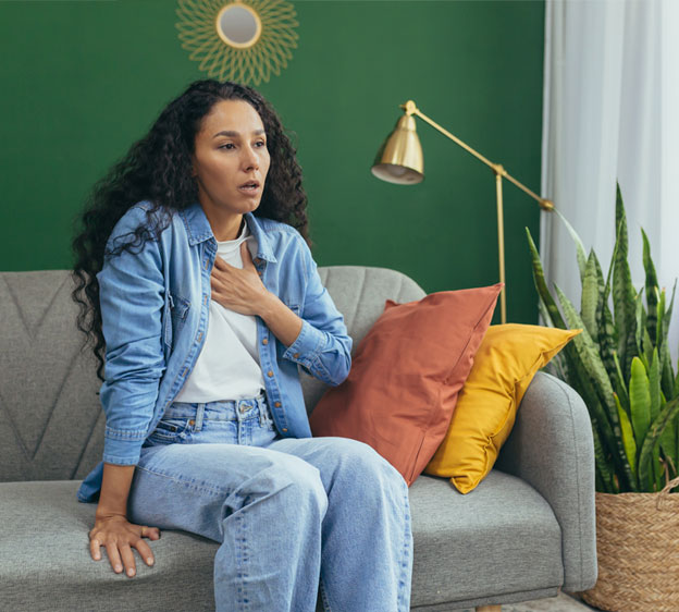 A middle-aged woman sits on a gray couch with her hand on her chest.