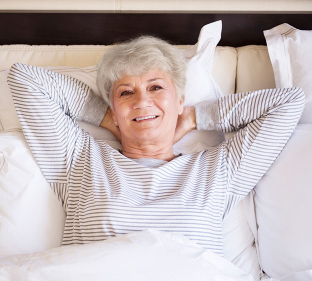 Older woman smiling and lying in bed with her arms around the back of her head