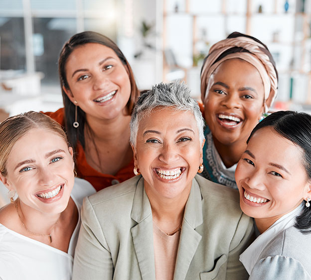 Five women of different ages and ethnicities stand in a group and smile at the camera.