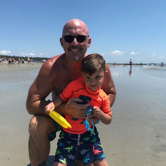 Beaufort Memorial Pharmacy Services Director Tim McCall poses for a photo on a beach with a young boy holding goggles and a beach toy.