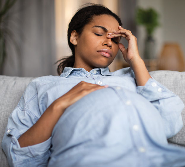 Woman in chambray shirt sitting on couch with her eyes closed and her left hand touching her forehead