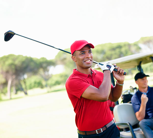 Two men on the golf course, one in the foreground wearing a red polo shirt and hat while holding a golf club over his shoulder, and the other sitting in a golf cart