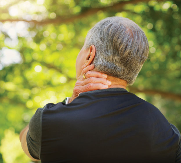 Older man rubbing the back of his neck with his hand with trees in the background.