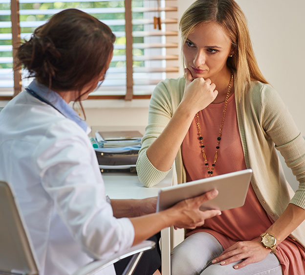 A younger woman in a cardigan sitting and speaking with a health care provider who is showing the woman a tablet.