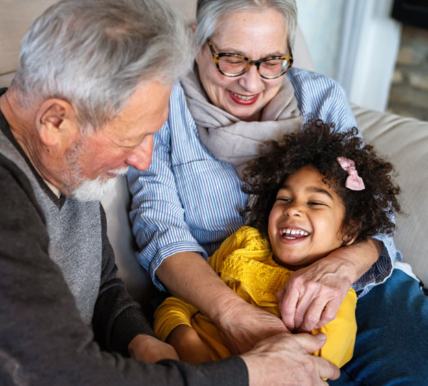 A little girl lays in her grandmother&rsquo;s lap laughing. Her grandmother is hugging her, and her grandfather sits next to them smiling.