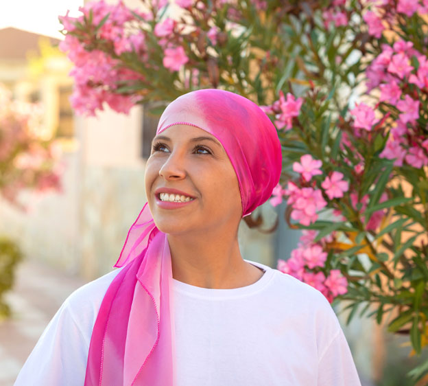 A woman wears a pink headscarf and stands in front of a tree blooming with pink flowers.