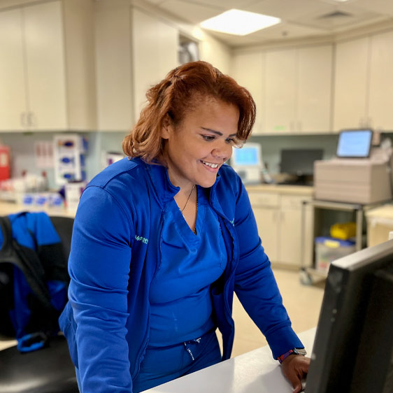 Meridith Godinez Carrillo stands at a nursing station in front of a computer.